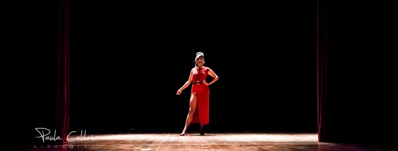 Foto da Drag Ashilleyy em um vestido lindo, vermelho, perna esquerda de fora. Ela está ao centro, em um palco de teatro muito iluminado. Canto esquerdo inferior está o credito da foto: Paula Caldas 'videeverso'.